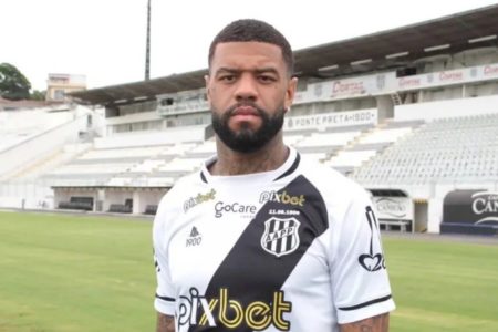 Imagem colorida mostra o lateral-esquerdo Junior Tavares, um homem negro, de barba, vestido com a camisa da Ponte Preta, que é branca com uma faixa transversal preta - Metrópoles