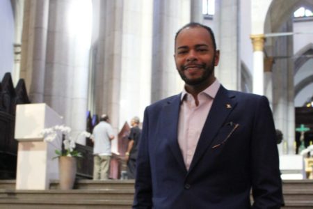 Imagem colorida mostra Fernando Alfredo, homem negro, de cabelos pretos e barba aparada, da cintura para cima, vestindo terno azul e camisa rosa, à frente da escadaria do altar da igreja da Sé - Metrópoles