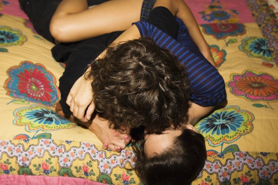 Foto colorida de dois jovens deitados em uma cama com lençol colorido abraçados e se beijando - Metrópoles