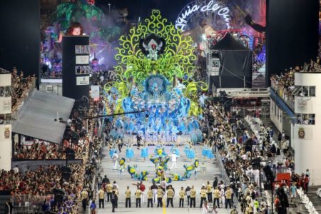 Imagem colorida mostra um dos desfiles de escola de samba em São Paulo, com alas em primeiro plano e um carro alegórico atrás - Metrópoles