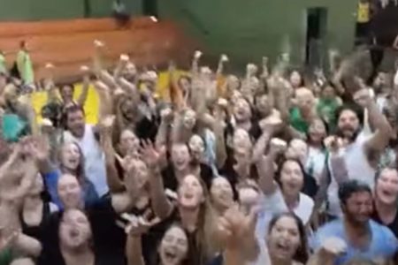 Imagem colorida mostra estudantes cantando hino de faculdade