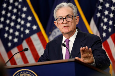 Imagem de Jerome Powell, presidente do Federal Reserve. Ele veste um terno azul escuro, camisa branca, gravata roxa e tem cabelos grisalhos e óculos. Ao fundo, duas bandeiras dos Estados Unidos - Metrópoles