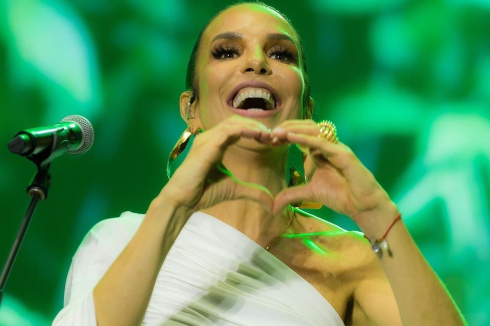 Ivete Sangalo posa no palco, com um look todo branco, fazendo coração para o público - Metrópoles