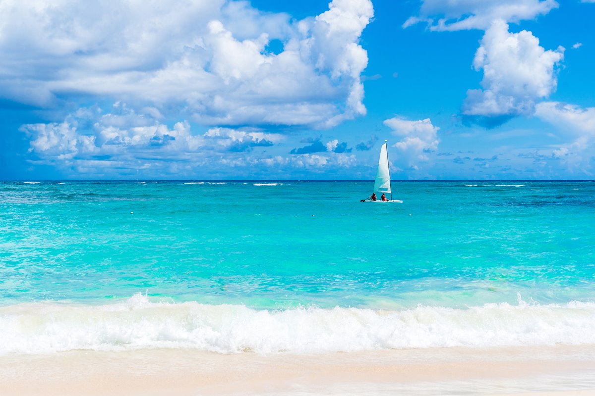 Foto colorida de praia de cor azul turquesa, com barco velejando - Metrópoles