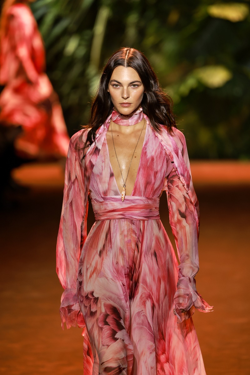 Foto colorida. Modelo branca, de olhos verdes e cabelos pretos soltos desfila na passarela com um vestido longo rosa - Metrópoles