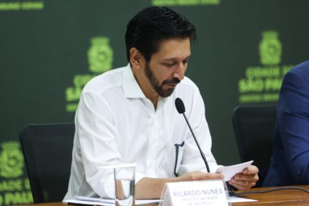 imagem colorida mostra homem branco, com barba e cabelo pretos. ele veste camisa branca, está sentado e lê um papel em frente a um microfone. metrópoles