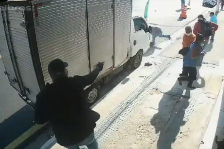 Imagem colorida mostra um bandido de blusa preta atirando em um PM à paisana que está correndo para se esconder na frente de um caminhão estacionado. Ele é observado por outro bandido de blusa vermelha - Metrópoles