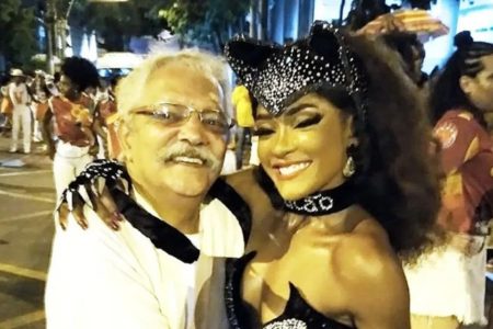 Foto de um homem branco, com cabelo grisalho e bigode grisalho e óculos de grau, abraçado com uma mulher preta fantasiada de mulher-gato - Metrópoles