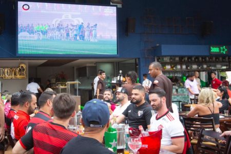 imagem colorida. Torcedores de costas assistindo ao jogo em uma tv de bar