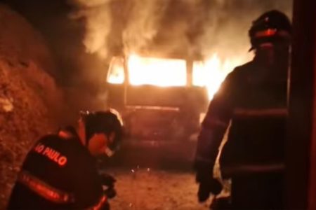 Imagem colorida de incêndio na Kombi. Há dois bombeiros na frente e o carro, em chamas, atrás - Metrópoles