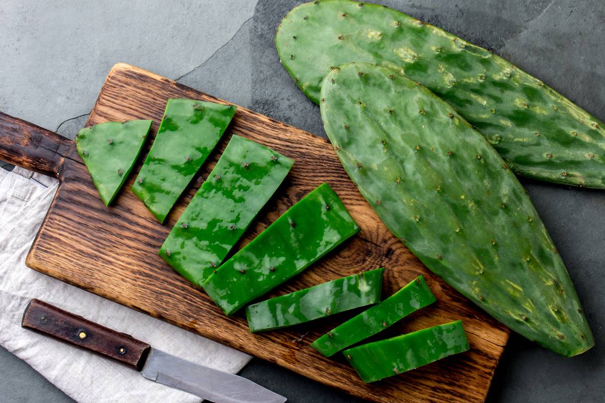 Foto colorida de cacto nopal, sendo um cortado em fatias e outro inteiro, próximo a uma tábua de madeira - Metrópoles