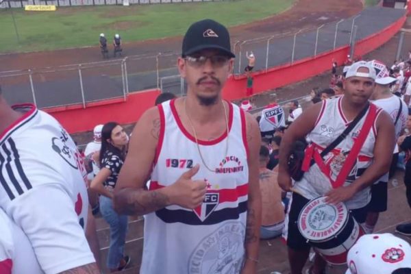 Foto colorida mostra homem com camisa regata do São Paulo, óculos de sol, boné e fazendo um 