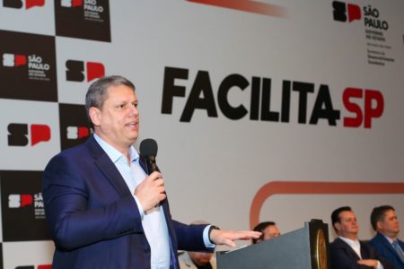 Imagem colorida mostra Tarcísio de Freitas, homem branco, grisalho, de terno azul e camisa branca, falando ao microfone em um palco com um banner do governo ao fundo - Metrópoles