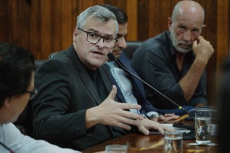 Imagem colorida mostra Rinaldi Digilio, homem branco, de cabelos brancos e óculos preto, vestindo roupas pretas, falando ao microfone enquanto gesticula com as mãos - Metrópoles