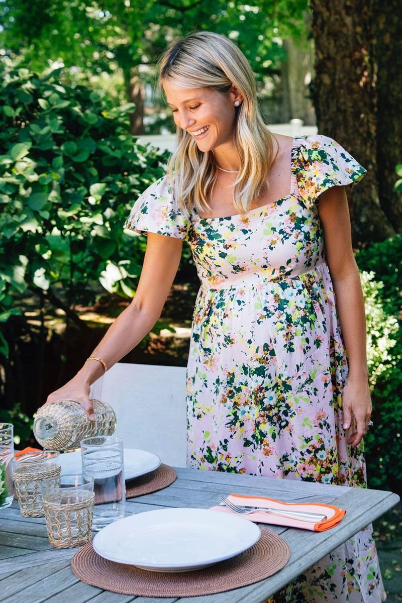 Foto colorida de mulher branca, loira e com vestido florido. Ela coloca água em um copo - Metrópoles