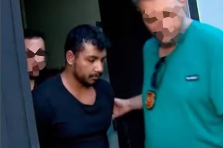 Em imagem colorida jovem de bigode e barba falha usa camiseta preta enquanto e conduzido por policial de camiseta azul água e distintivo no peito - Metrópoles