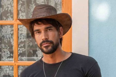 Foto colorida do ator Matteus Cardoso. Ele usa chapéu marrom, uma blusa preta e um cordão prata. É um homem branco de barba e cabelo preto - Metrópoles