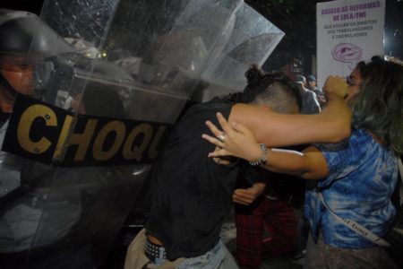 Em foto colorida PM com escudo no qual está escrito Choque empurra jovem que bate braço em rosto de mulher