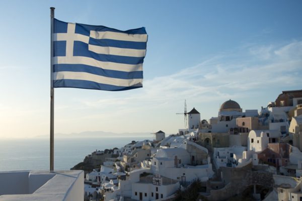 Imagem colorida da bandeira da Grécia, em destaque, tremulando em uma dia de céu azul. Ao fundo, o mar e um conjunto de casas em um cenário paradisíaco - Metrópoles