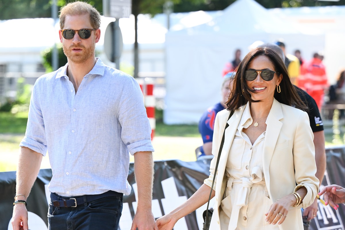 Harry e Meghan aparecem na imagem de mãos dadas. Ambos usam roupas claras e óculos escuros.