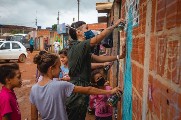 Mulher de máscara grafitando em parede de tijolos. Acompanhada por crianças - Metrópoles