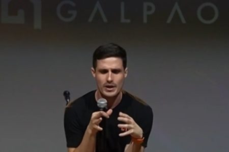 Em foto colorida homem branco de camiseta preta fala do microfone - Metrópoles