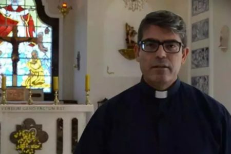 Imagem colorida mostra o padre Ferdinando Henrique Paiva Rubio, um homem branco, de cabelos grisalhos, vestido com uma batina preta ao lado de um altar religioso dentro de uma Igreja - Metrópoles