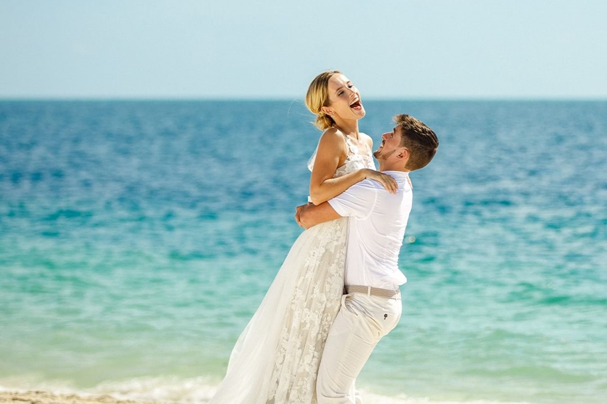 Foto colorida de mulher e homem, com roupa de noivos, próximo ao mar. O homem segura a mulher no colo - Metrópoles