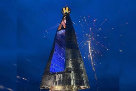 Imagem colorida de monumento de Nossa Senhora de Aparecida, coberto por luzes azuis e amarelas - Metrópoles