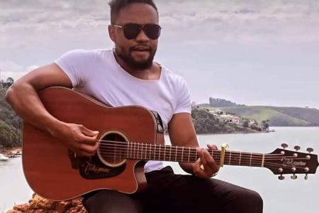 Foto colorida de homem de óculos escuros segurando violão, em frente ao mar; cantor é suspeito de assassinar dentista - Metrópoles