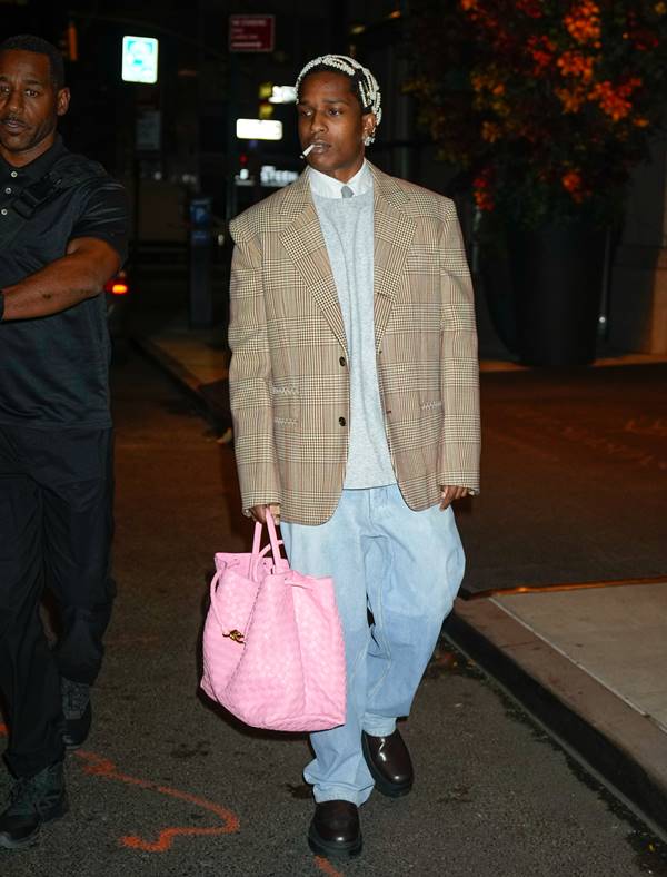 A$ap Rocky está entre os homens que usam bolsas A$ap Rocky usando look com calça jeans e blazer cinza oversized. Ele segura uma bolsa rosa e tem um cigarro na boca - Metrópoles