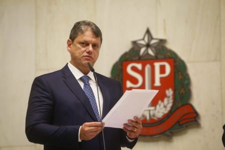 Fotografia colorida mostra Tarcísio de Freitas segurando uma folha de papel sulfite enquanto olha para o horizonte posicionado em frente a um microfone - Metrópoles