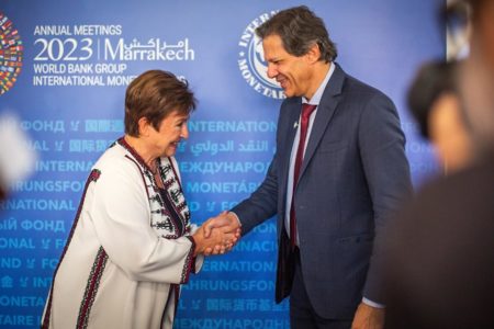 Fernando Haddad e a diretora-geral do FMI, Kristalina Georgieva, se cumprimentam. Ele veste um terno azul, e ela, uma blusa branca com detalhes coloridos - Metrópoles