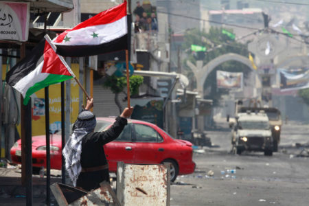 Imagem colorida de um homem segurando as bandeias da Palestina e da Síria - Metrópoles