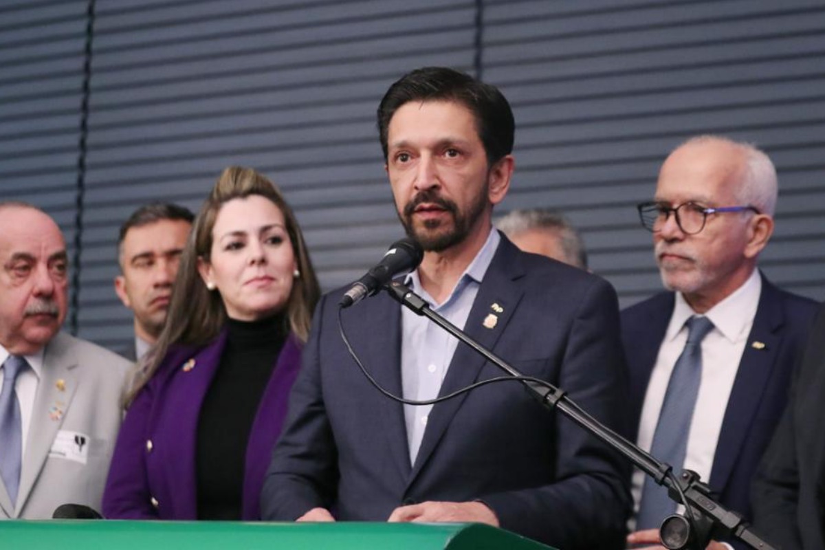 Imagem mostra Ricardo Nunes, homem branco de cabelo e barba pretos, de terno cinza escuro e camisa cinza clara, falando ao microfone, no centro de um grupo de pessoas - Metrópoles