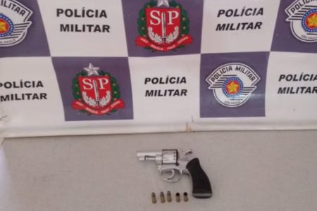 Foto colorida mostra arma de calibre 32 ao lado de cinco munições. Ao fundo, é possível ver um banner da Polícia Militar