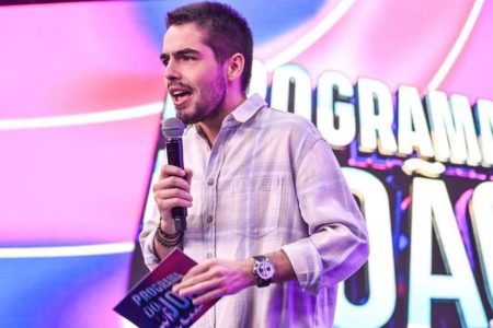 Foto colorida de João Guilherme Silva. Ele é um homem branco, cabelo e barba preta, usa blusa clara e quadriculada. Está em um palco falando no microfone - Metrópoles