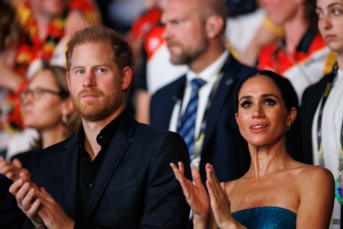 Harry, homem branco e ruivo está usando um terno todo preto. Meghan, uma mulher negra, está com os cabelos amarrados em coque e usando um vestido de gala azul. Ambos estão aplaudindo - Metrópoles