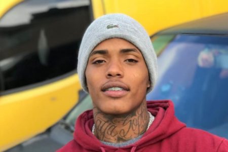 Imagem colorida mostra o rapper LK Metralha, um homem negro, magro, com tatuagens no pescoço, vestido com um gorro cinza e uma blusa vermelha - Metrópoles