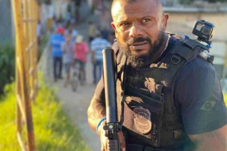 Imagem colorida do Delegado Da Cunha. Ele é um homem negro, careca e de barba e, na foto, aparece de uniforme da Polícia Civil, segunrado uma arma