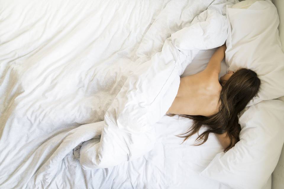 Foto colorida de uma mulher de cabelos castanhos deitada de bruços em uma cama branca sem roupa - Metrópoles