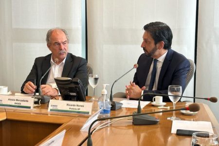 Imagem colorida mostra mesa de reuniões com Aloisio Mercadante, homem branco, calvo, de bigode, terno cinza escuro e camisa branca, conversando com Ricardo Nunes, homem branco, de cabelo e barba pretos, de terno e gravata azuis - Metrópoles