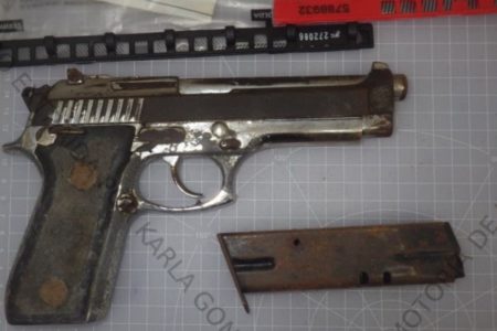 Imagem colorida de pistola calibre 9 milímetros. Com marcas de ferrugem, arma está de lado, sobre uma superfície, com o cano para a direita.