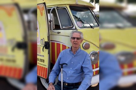 homem idoso branco de camisa social azul sentado olhando para frente. Atrás há a van do Churros do Tio