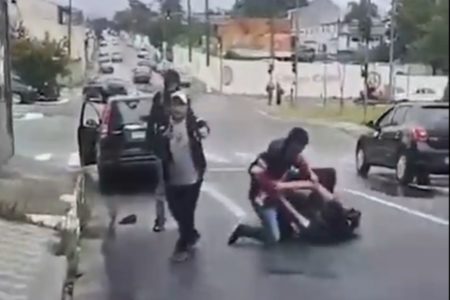 Imagem colorida mostra dois adolescentes brigando amontoados no chão enquanto um homem segura uma arma