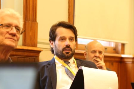 Imagem colorida mostra Guilherme Piai, homem branco, jovem, de cabelo e barba pretos, vestindo terno azul, camisa branca e gravata cinza, sentado ao lado de homens grisalhos, em uma sala com paredes amarelas - Metrópoles