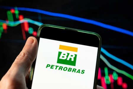 Telefone celular com a imagem do logotipo da Petrobras. Ao fundo, painel com o movimento de ações na Bolsa de Valores - Metrópoles