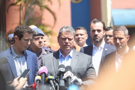 Imagem colorida mostra Tarcísio de Freitas, homem branco, grisalho, de terno cinza e camisa azul, cercado de auxiliares, de frente para microfones - Metrópoles