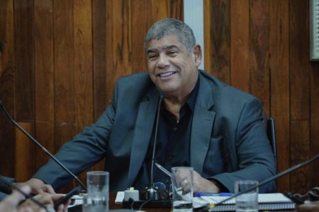 Imagem colorida mostra Milton Leite, homem negro, de terno cinza e camisa preta, sentado em uma mesa de reuniões em uma sala com paredes de madeira. Ele sorri para sua direita - Metrópoles