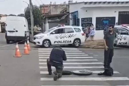 Imagem colorida de rua em que corpo foi encontrado pela Polícia. Foto mostra homem agachado em bueiro, com uma viatura da PM atrás - Metrópoles